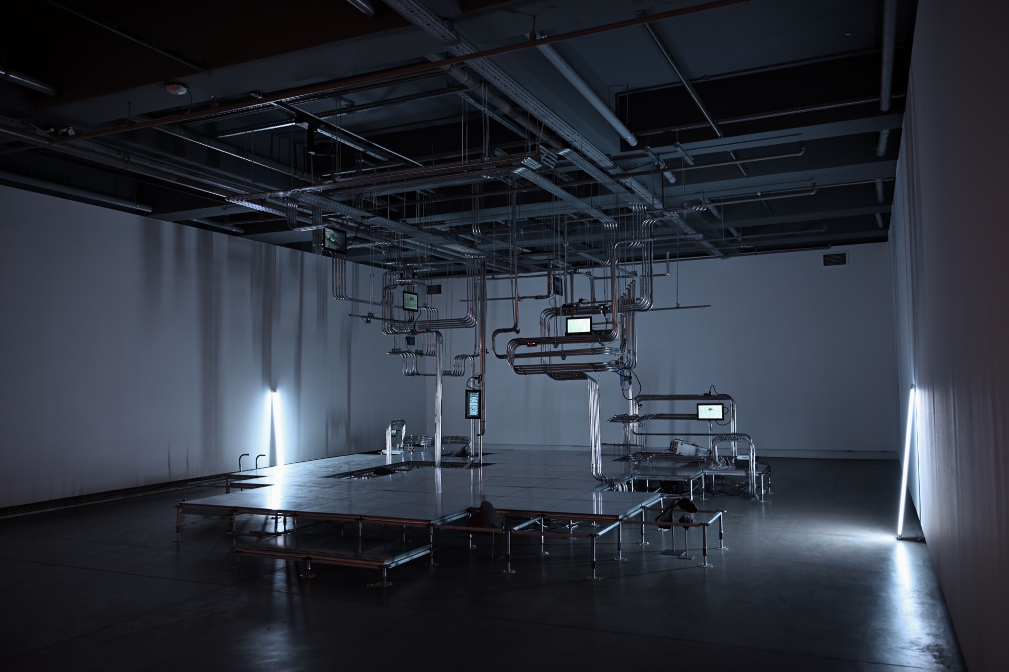 Installation view: suspended metal pipe structure with small screens over a raised platform, lit by vertical lights along the walls
