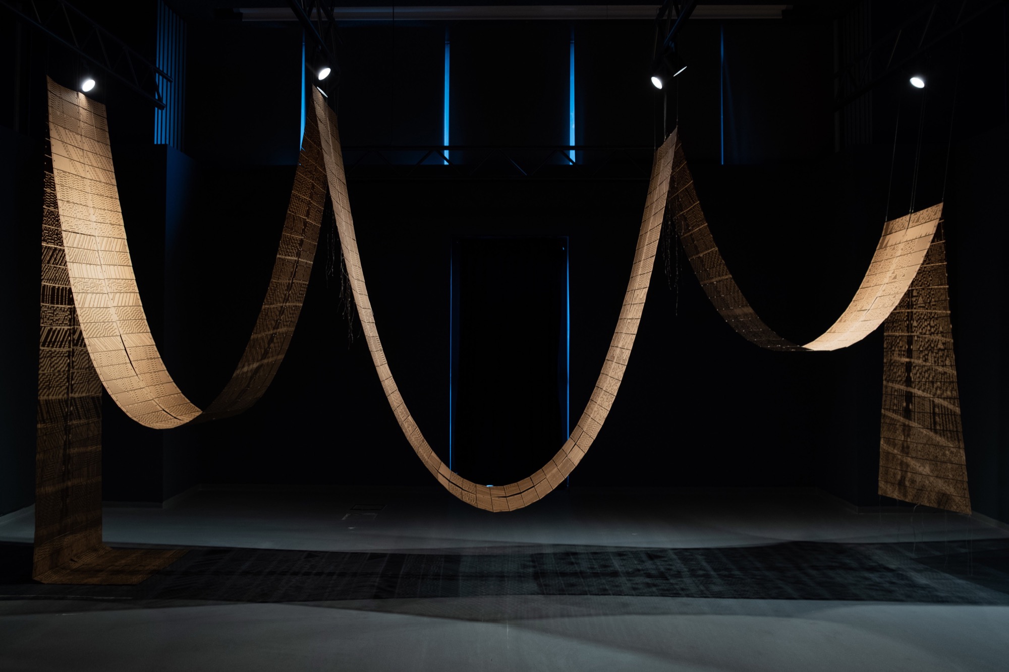 Installation view: long perforated panel strips suspended in sweeping curves across a dark space