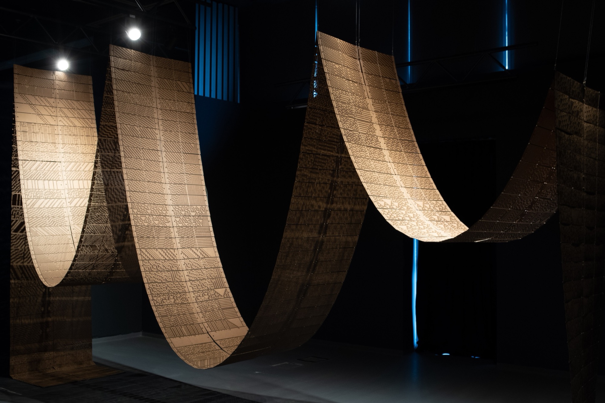 Installation view: perforated strips hanging in loops from the ceiling, lit by spotlights