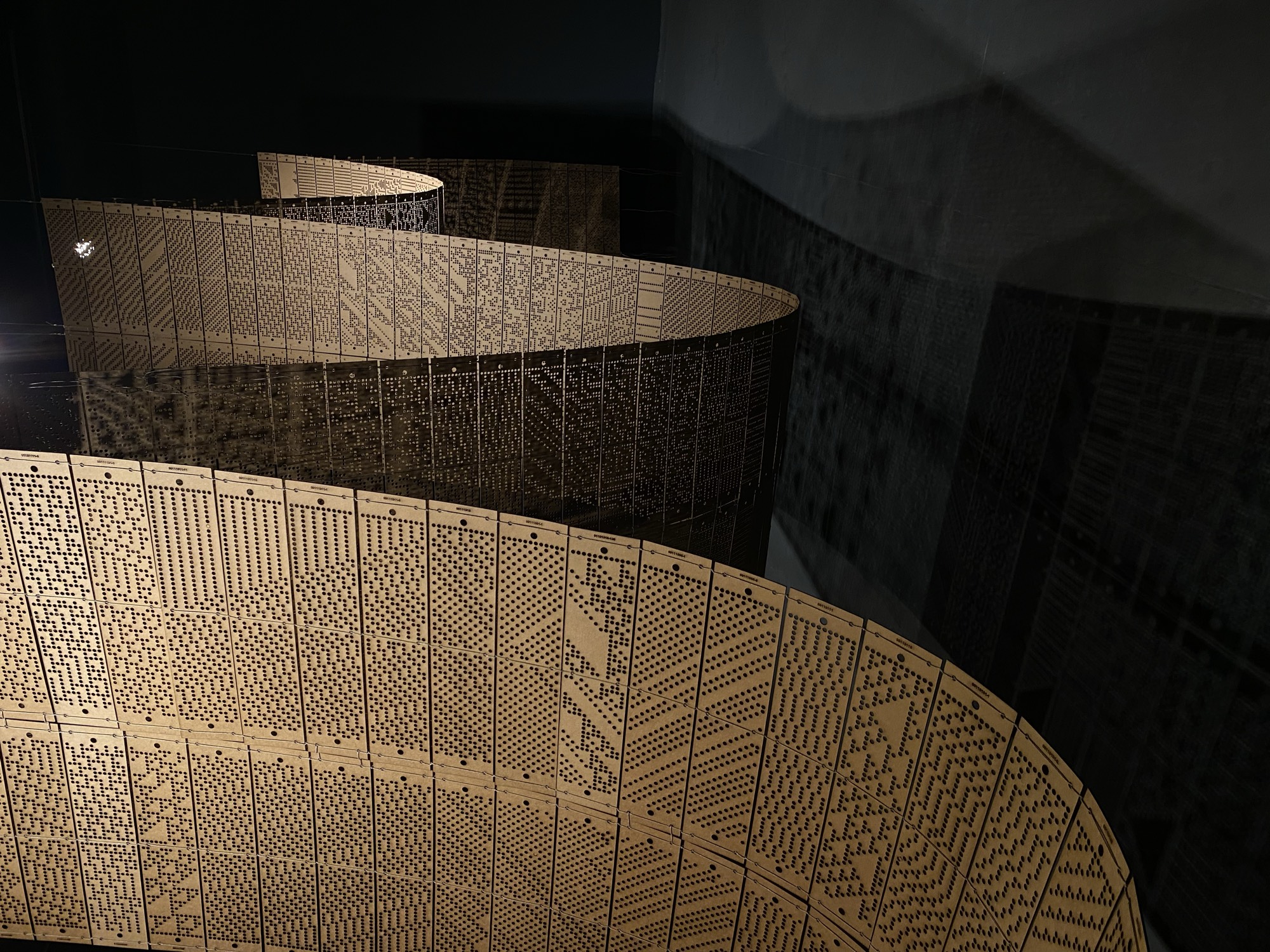Installation view: tall perforated panel strips hanging in deep curves above a dark floor, their dot patterns casting shadows