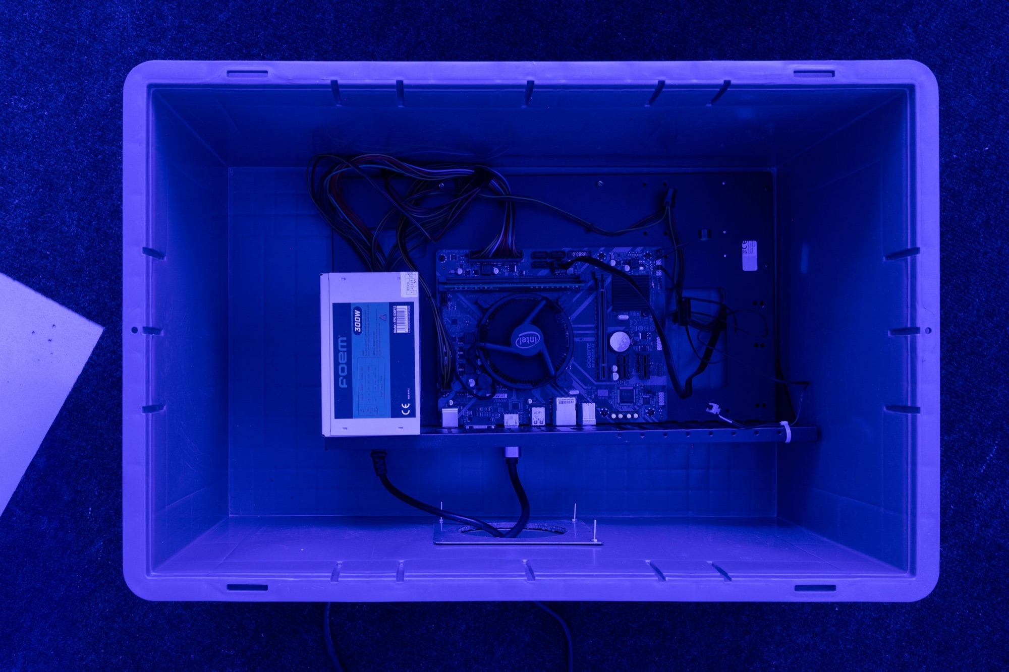 Top view of an open plastic crate under blue light, containing a computer motherboard, cables and a hard drive