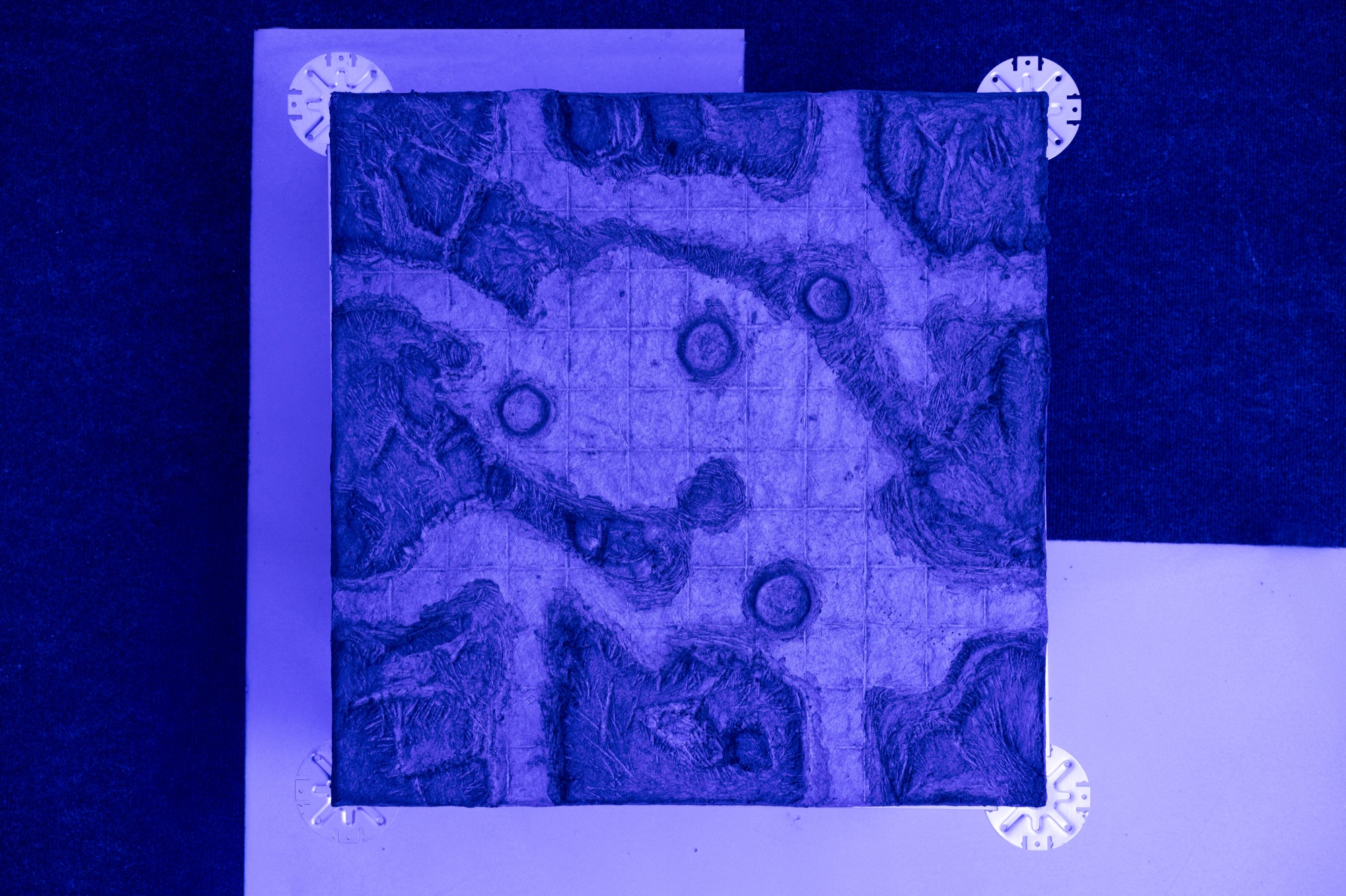 Top view under blue light: a square panel with a carved, textured relief of channels and small round craters over a faint grid, resting on a raised floor support with metal pedestal plates at its corners