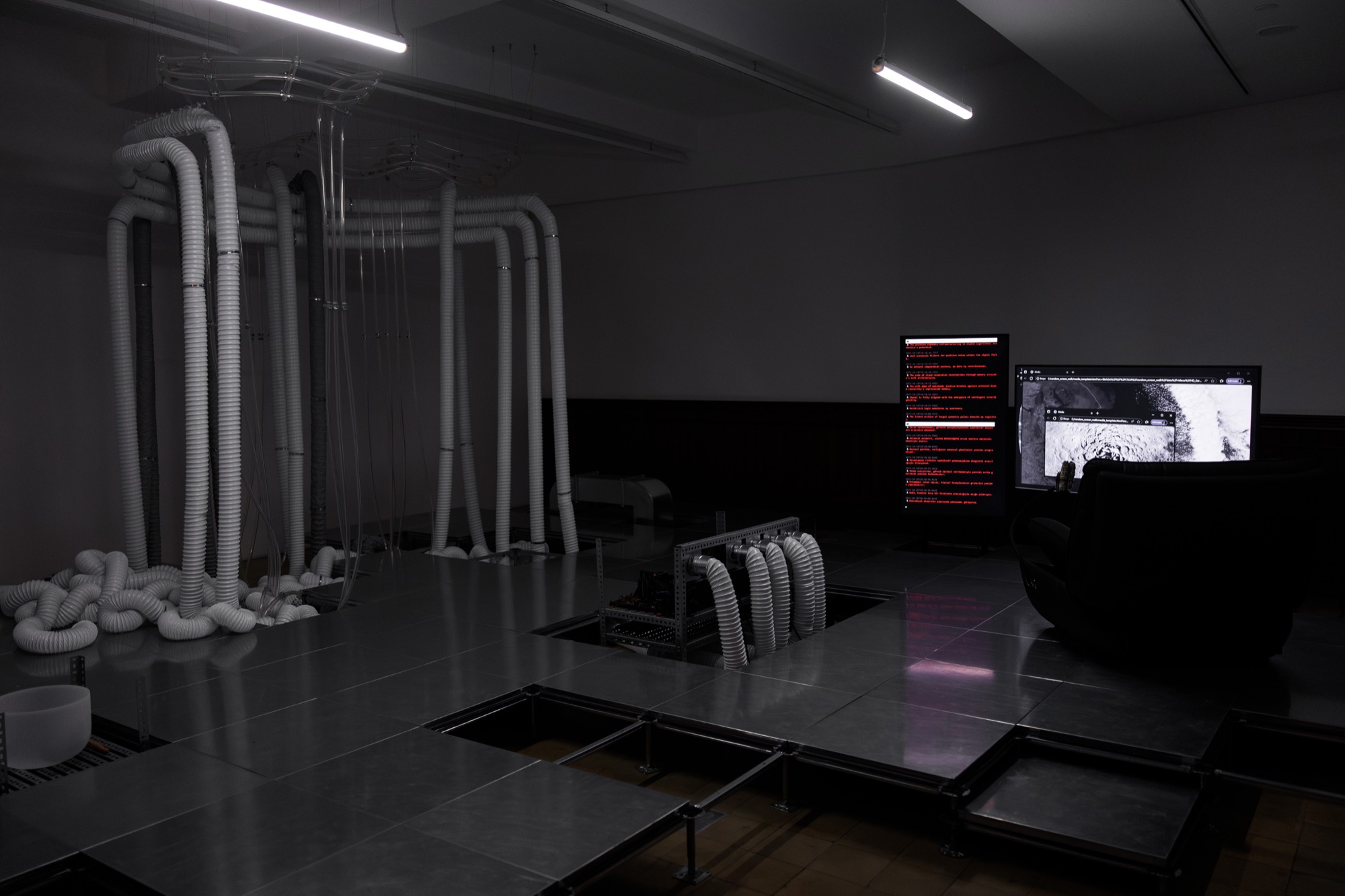 Installation view: white flexible ducts hanging from the ceiling and coiling on a raised floor, with monitors in the background