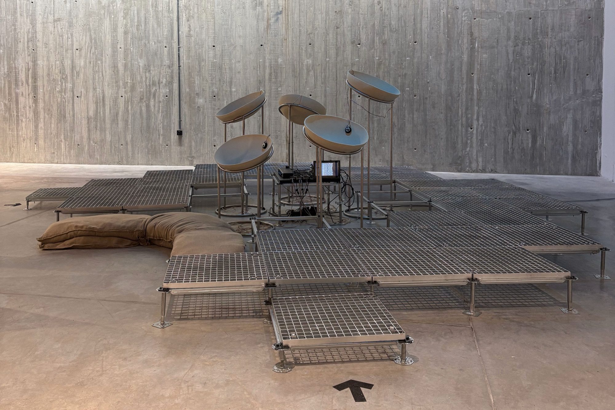 Installation view: dish-shaped sound elements on metal stands above a raised floor of steel grating panels, with sandbags, in a concrete-walled gallery