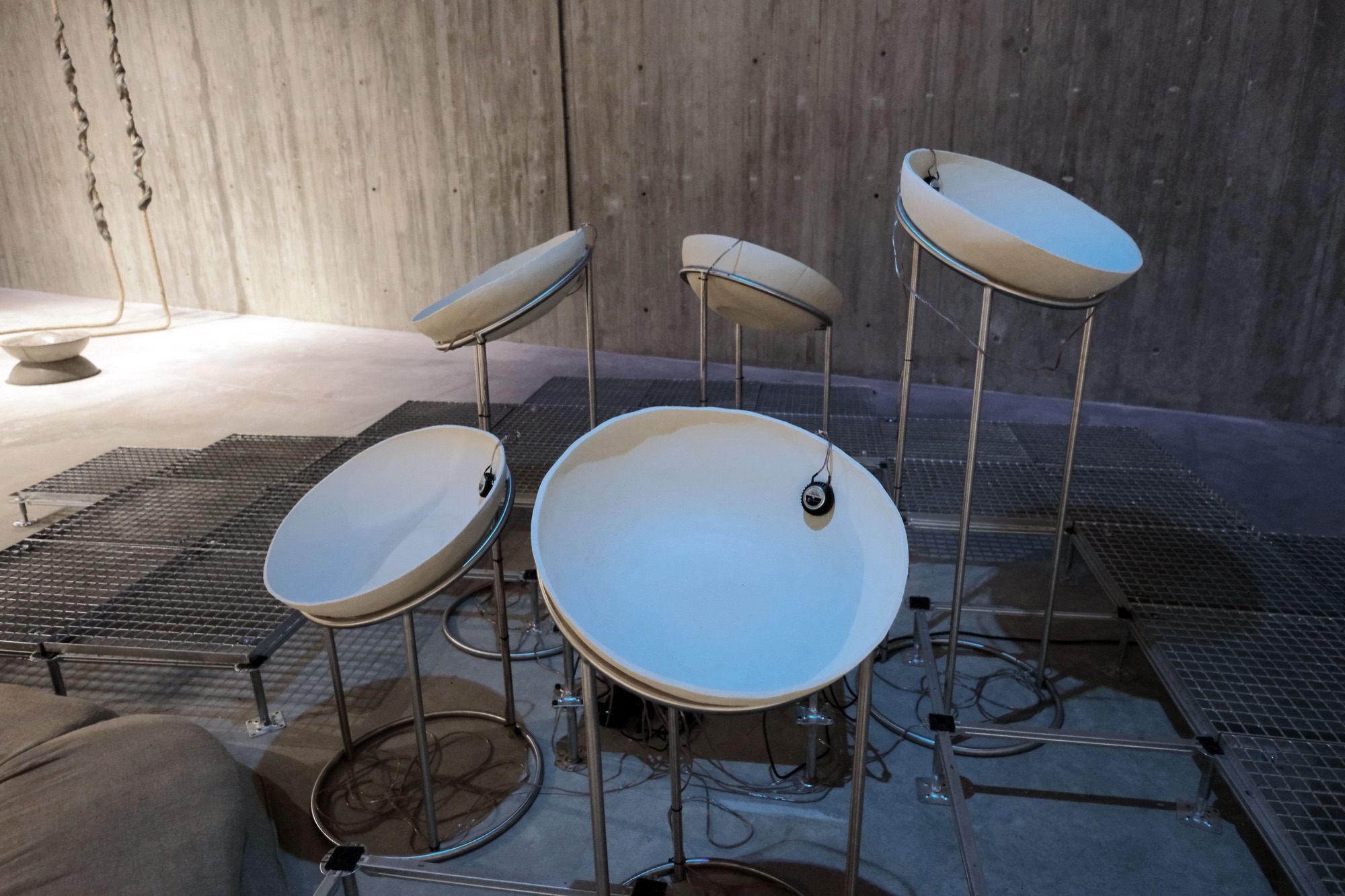 Detail: dish-shaped sound elements with small speakers attached to their rims, on steel stands over a raised grating floor, a concrete wall behind