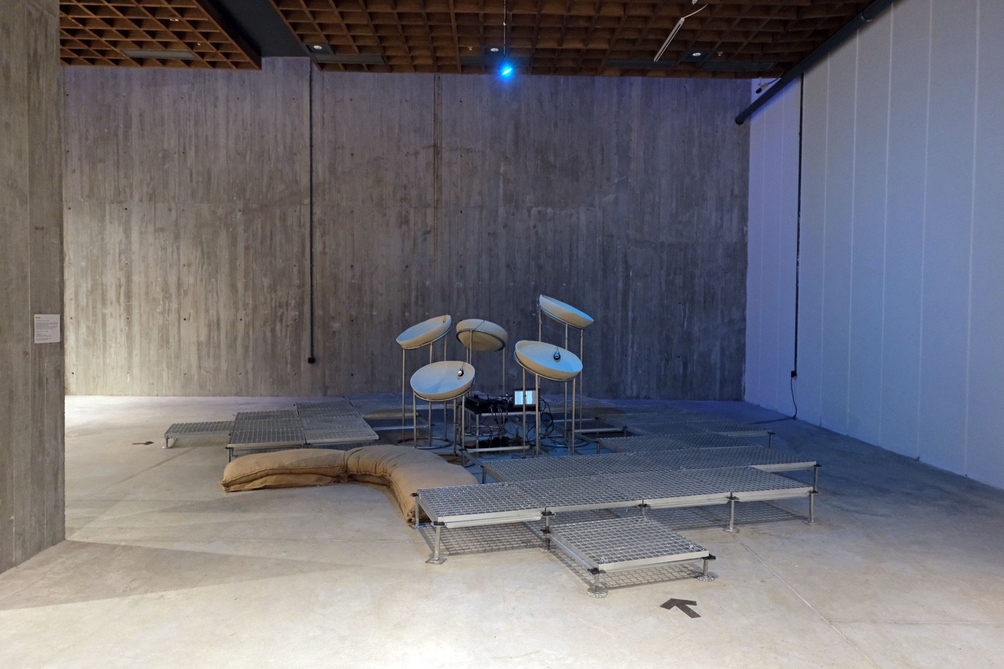 Wide installation view in a concrete hall: the cluster of dish-shaped sound elements stands on a low platform of steel grating panels, with a curved line of sandbags along its edge